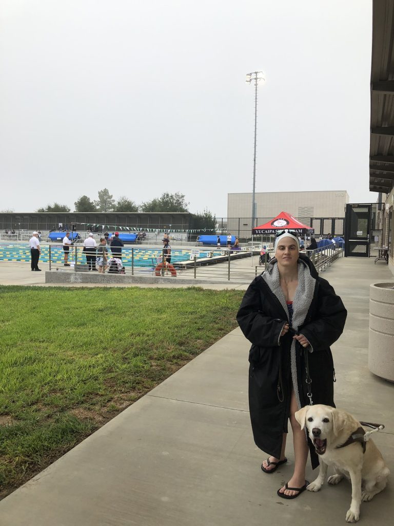 Megan stands at the edge of a competitive swim meet wearing a black hooded swim parka and a white swim cap. Suri, her yellow Labrador guide dog, sits next to her in working harness. The pool, lane judges, and stadium lights are visible behind them.
